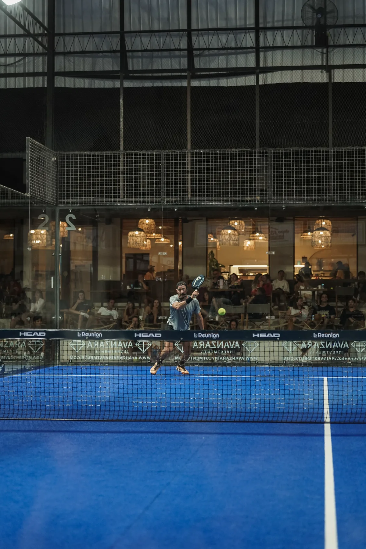 An evening match on a floodlit blue padel court, spectators watching from the lit clubhouse behind the glass